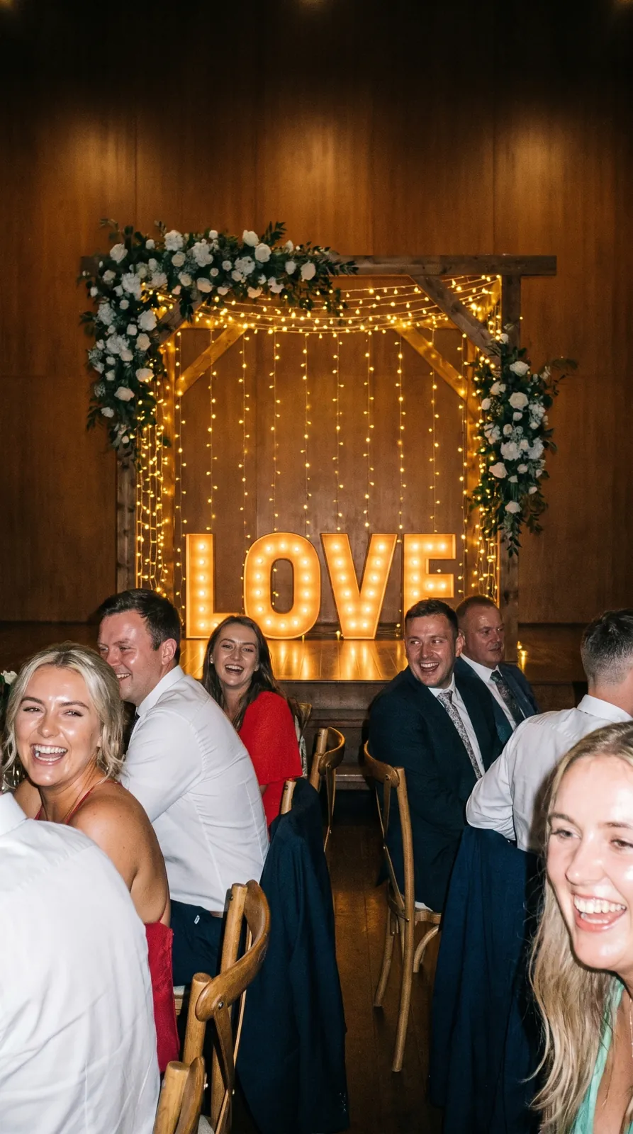 Gasten poseren bij LOVE-lichtletters met bloemenboog op een bruiloft